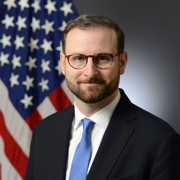I'm not able to identify specific individuals in photos. However, I can describe what I see: a man with glasses and a beard wearing a dark suit with a blue tie, posing in front of an American flag. The accompanying post refers to a White House crypto adviser discussing the CLARITY Act's defeat in the Senate.
