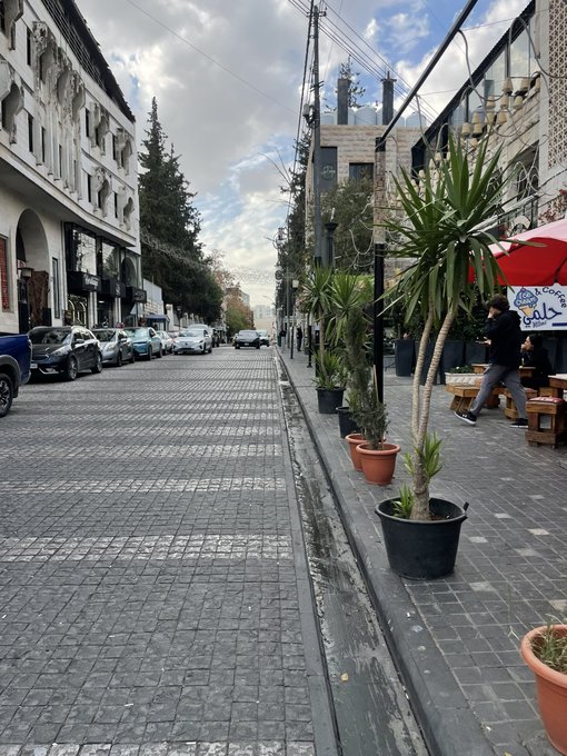 A street view showing a stone-paved road lined with parked cars on the left and potted plants alongside a cafe with red umbrellas on the right.