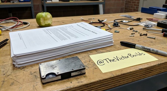A stack of papers sits on a wooden workbench next to a black Sharpie, screws, cables, and a yellow sticky note reading '@TheVictorBuilds'.