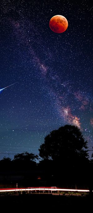 A night sky photograph captures a red blood moon and a bright shooting star streaking across the Milky Way above a silhouetted tree line and light trails on the ground.