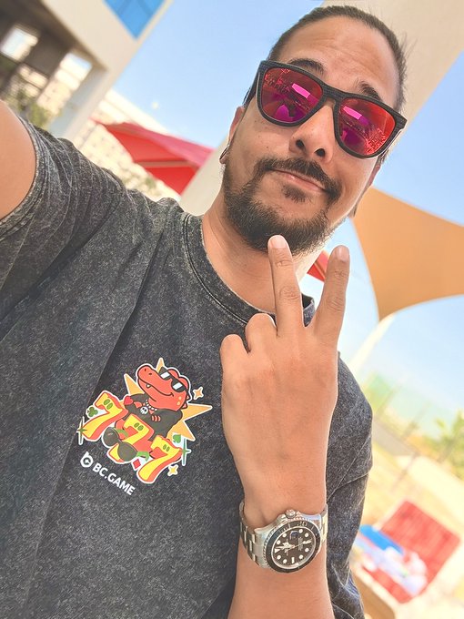 Selfie of a bearded man wearing red reflective sunglasses and a gray BC.GAME t-shirt with a dragon graphic, making a peace sign outdoors.