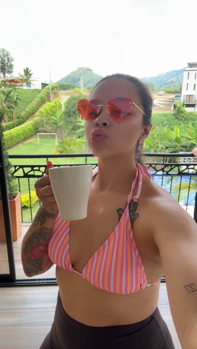 A woman wearing heart-shaped sunglasses and a striped pink and orange bikini top holds a white mug while posing on a balcony overlooking a green, hilly landscape.