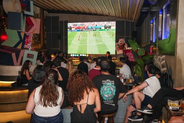A crowded bar or lounge where a group of people sit on tiered seating watching a tennis match on a large projection screen.