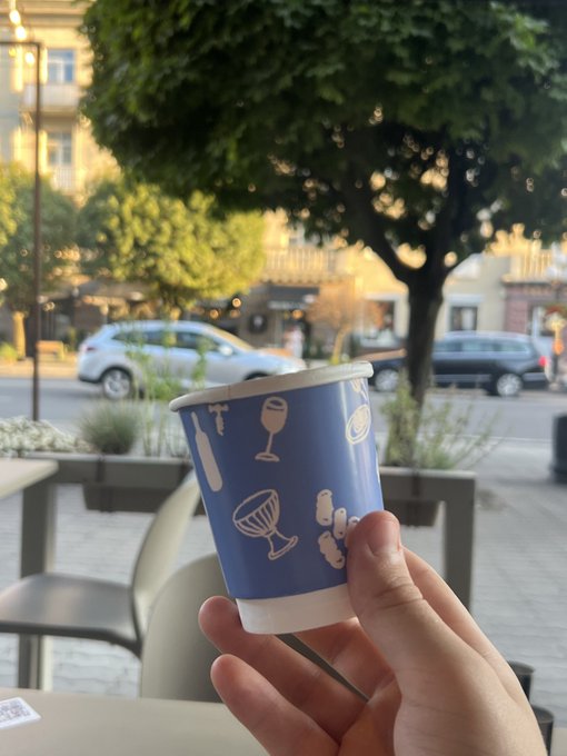 A hand holds a blue paper cup decorated with white icons of bottles, wine glasses, and grilling tools, with a blurred European street scene featuring parked cars and trees in the background.