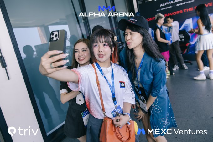 Three young women taking a selfie at the ALPHA ARENA event, with logos for triv and MEXC Ventures visible.