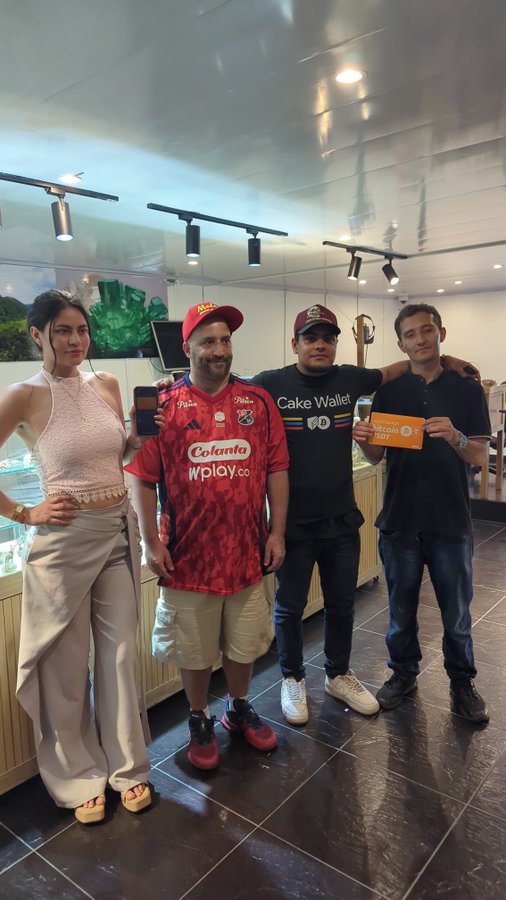 Four people posing together indoors at what appears to be a crypto event - a woman in a beige outfit, a man in a red "Colanta wplay.co" soccer jersey, a man in a "Cake Wallet" shirt and sunglasses, and a man holding an orange "Bitcoin NOW" sign.