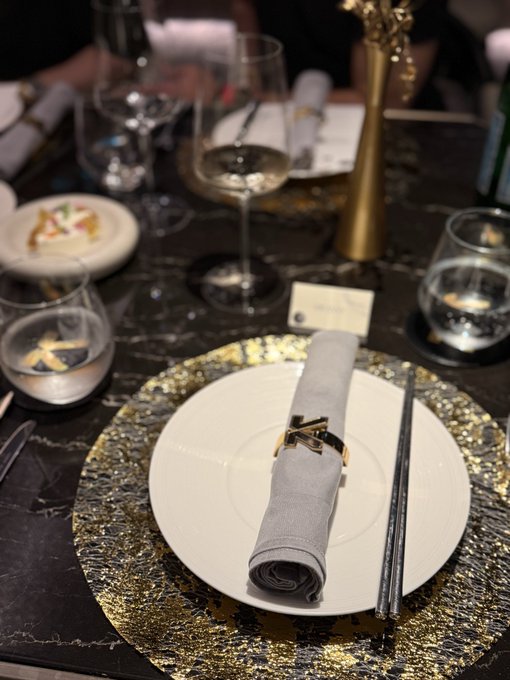 An elegantly set dining table featuring a white plate atop a textured gold charger, with a grey napkin secured by a decorative ring, accompanied by wine glasses and chopsticks.