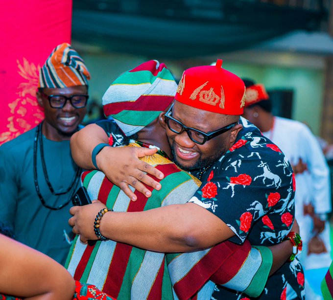 Two men in colorful traditional Nigerian attire embrace warmly, one wearing a striped cap and the other a red cap with a gold emblem, while a smiling man in a teal outfit and glasses watches in the background.