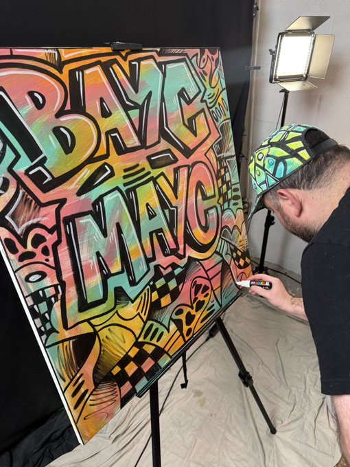 An artist wearing a colorful patterned cap is creating a vibrant graffiti-style artwork featuring the words 'BE BRAVE' and 'BE BANG' on an easel, illuminated by a studio light.