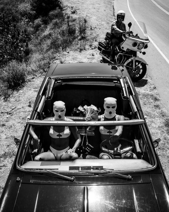 A black and white photograph shows two people in bikinis and ski masks sitting in a convertible car filled with money, while a police officer on a motorcycle watches from the road behind them.