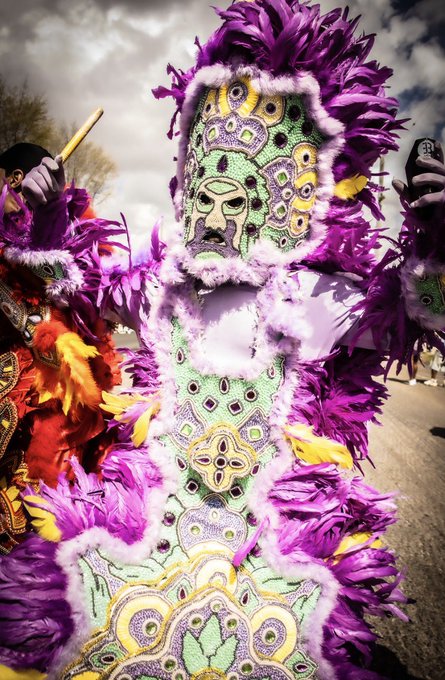 A vibrant Mardi Gras Indian in an elaborate costume featuring purple, yellow, and green feathers with intricate beaded patterns and a decorated mask, photographed outdoors.