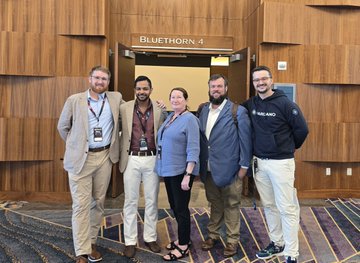 Four people (three men and one woman) posing together in a conference hallway with wood-paneled walls under a sign that reads 'BLUETHORN 4'.