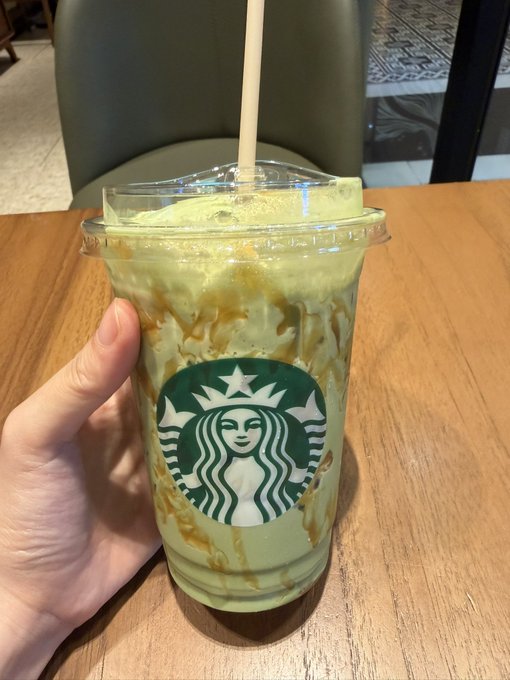 A hand holding a clear Starbucks plastic cup containing a green blended beverage with caramel drizzle.