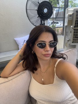 A selfie of a woman with dark hair wearing sunglasses, gold hoop earrings, and a white ribbed tank top, sitting outdoors near a fan.