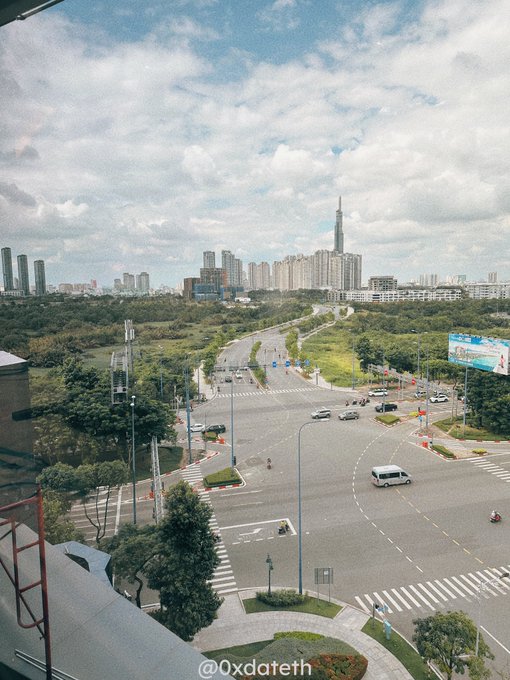 A high-angle view of a wide, multi-lane intersection in an urban Vietnamese city with the Landmark 81 tower visible in the distance, captured under a partly cloudy sky.

The watermark "@0xdateth" is visible at the bottom. The expansive cityscape and morning sky complement the post's "good morning, new day, let's get to work" sentiment, offering a fresh view of the city to start the day.