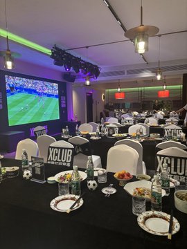 The image shows an XCLUB VIP event setup featuring rows of black-draped tables with white chair covers, soccer-themed decorations, and a large screen displaying a football match, where attendees are enjoying refreshments in a sports lounge atmosphere. The setup ties into the post's mention of OKX's immersive, perk-filled events, with the XCLUB branding and football broadcast reflecting the lively, on-site entertainment experience described.