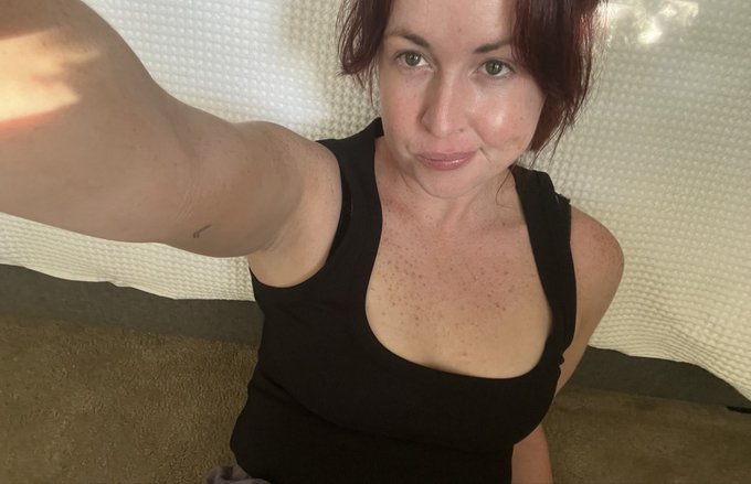 A selfie of a woman with dark red hair wearing a black tank top, taken from a slightly low angle indoors.