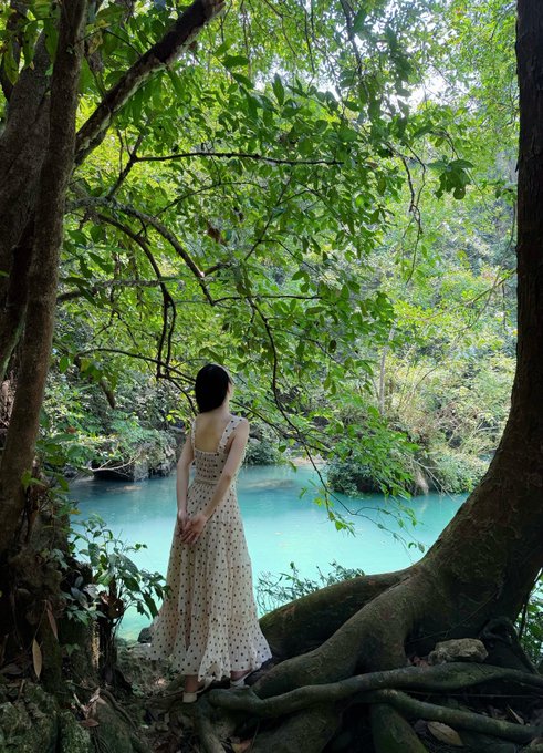 A woman in a white polka-dot sundress stands on tree roots looking out at a vibrant turquoise river surrounded by lush green forest foliage.