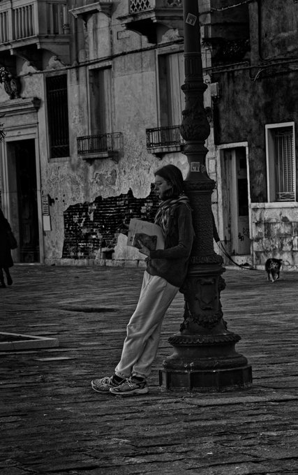 urns on a balcony, a person reading a book, and a dog walking by are present in the black and white photo of a city street corner.