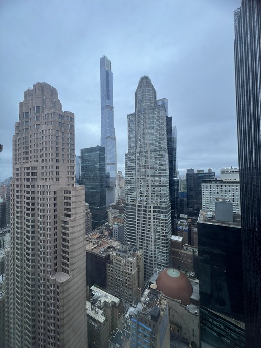 A high-angle photograph of a dense city skyline under an overcast sky, featuring a prominent tall skyscraper with a spire rising above other modern buildings, including one with a distinctive copper-colored dome.
