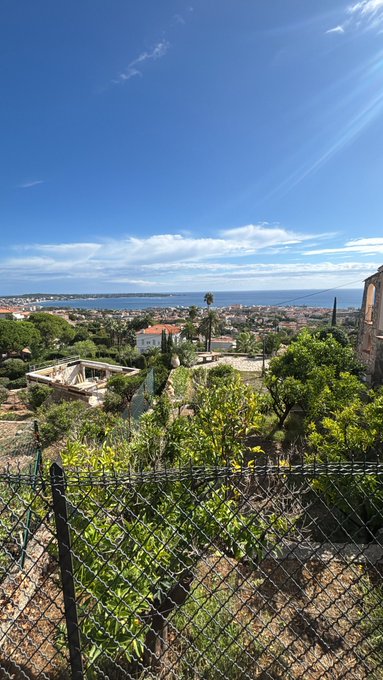 A scenic view from an elevated vantage point shows a Mediterranean coastal town with red-tiled roofs, palm trees, and the sea in the distance under a bright blue sky with wispy clouds.
