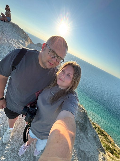A selfie of a couple on a cliff overlooking the ocean with the sun shining brightly; the man wears a grey shirt and glasses with a camera strapped across him, and the woman wears a matching grey shirt and white shorts.