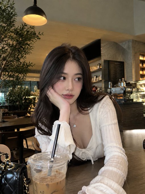 A young woman with long dark hair poses at a café, resting her chin on her hand while wearing a white knit top, with an iced coffee drink on the table in front of her.