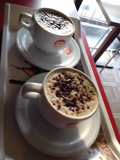 Two cups of frothy cappuccinos topped with chocolate powder are served on white saucers on a tray.