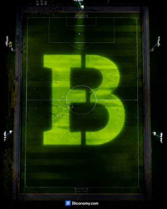 The image features a large soccer field with the word "B" written across it, which appears to be illuminated by green lights. The photo was taken from an aerial perspective, providing a unique view of the field's layout and lighting conditions. The caption at the bottom of the image reads "Biconomy fam! The World Cup is kicking off, and so is your chance to score BIG! Believe in your picks, trust the game, and watch your predictions pay off". This message seems to be related to the soccer event captured in the photo, as it encourages viewers to believe in their choices and trust the outcome of the game.