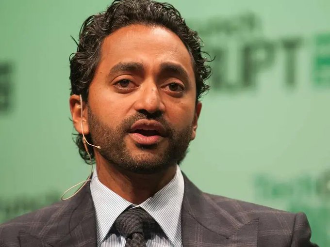 A close-up portrait of a bearded man in a dark plaid suit and tie, wearing a headset microphone, speaking against a blurred green background with partial text visible.