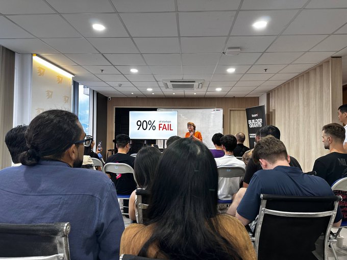 A photograph taken from the back of an audience attending a presentation, with a screen displaying '90% of Startups FAIL' while a speaker holds a microphone and a 'BUILDER GRANTS' banner stands to the side.