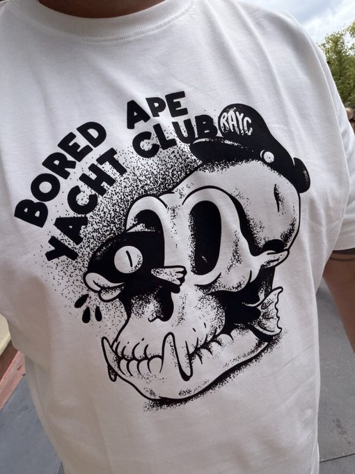 The image features an individual wearing a white t-shirt with a graphic design of a skull and hat, labeled "Bored at a Pirate Club". The person is standing near a building, likely taking a break from exploring or walking around Epcot. In the surrounding environment, there are trees visible in the background, adding to the theme of a pirate-themed location.