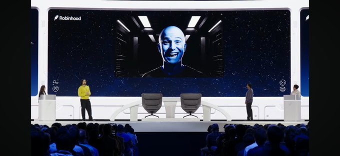 The image shows a Robinhood Hood Summit stage with Vladimir Tenev appearing via video call on a large screen, smiling broadly while being introduced to a live audience of attendees. The caption references Tenev taking playful jabs at Coinbase's Brian Armstrong and the Base network during the event, suggesting a lighthearted industry rivalry moment.