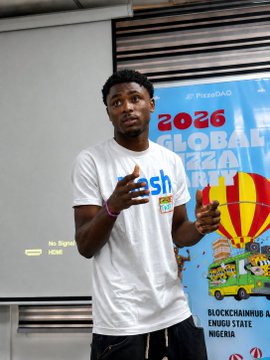 A young man in a white t-shirt with 'flash' written on it speaks while gesturing, standing in front of a banner for the '2026 GLOBAL ENTREPRENEURSHIP WEEK' in Nigeria.