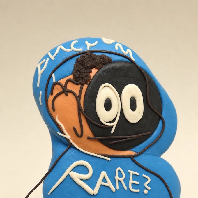 A blue clay sculpture featuring an orange face with large cartoon eyes, topped with dark curly hair, displaying the white text 'ANONYMOUS' and 'RARE?' on its body.