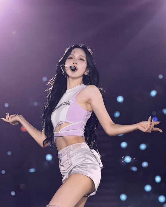 A female performer with long dark hair, wearing a pink and white crop top and white shorts, performing on stage with a headset microphone.