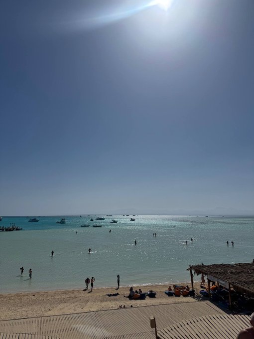 A sunny beach scene with people swimming in clear turquoise water, boats anchored offshore, and a wooden deck with a thatched-roof structure in the foreground.