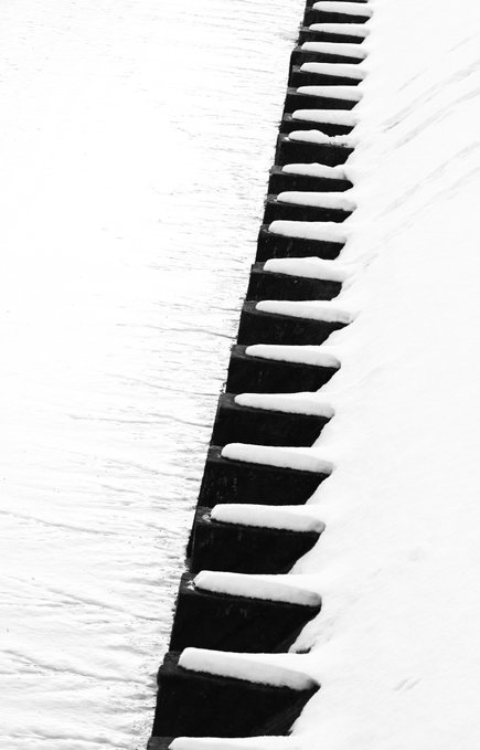 A black-and-white photograph showing a row of dark, rectangular blocks or steps, topped with snow, receding diagonally through a snow-covered surface.