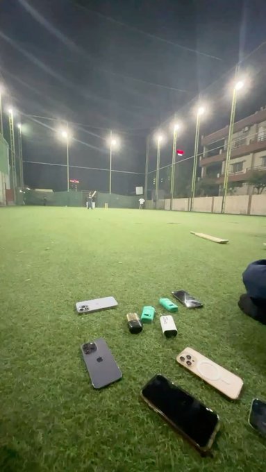 Caption: Several smartphones lie scattered on the turf of an indoor cricket facility at night, while players bat in the distance under bright stadium lights — illustrating a fun evening of indoor cricket with friends before returning to market analysis.