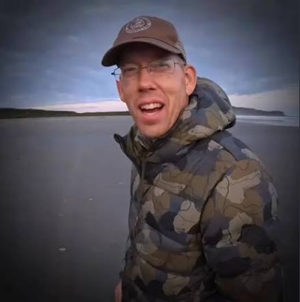 A man wearing glasses, a brown cap, and a camouflage jacket, smiling outdoors against a backdrop of a cloudy sky and water.