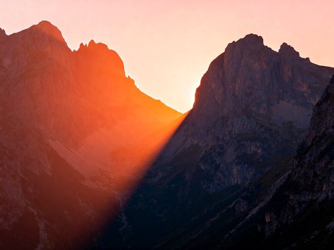 A scenic photograph of mountain peaks at sunset with vibrant orange and pink colors illuminating the sky and casting light over the rugged terrain.