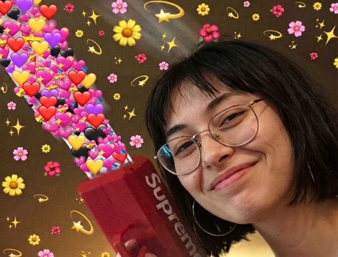 The image features a young woman with glasses holding up a red box that says "Supreme" on it. She has a big smile on her face as she poses for the photo. The background of the image is filled with various flowers, including roses and daisies, which adds to the overall visual appeal of the scene.