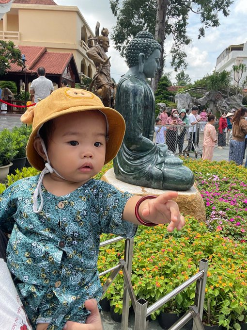 A young child wearing a yellow sun hat and a floral green shirt points toward a bronze Buddha statue surrounded by flowers and onlookers at what appears to be a temple.