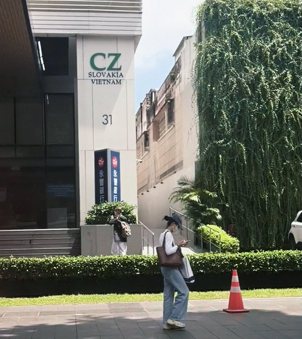 A street-level view of a building labeled 'CZ SLOVAKIA VIETNAM 31' with Bank of China signage, where a person wearing a mask looks at their phone near an orange traffic cone.