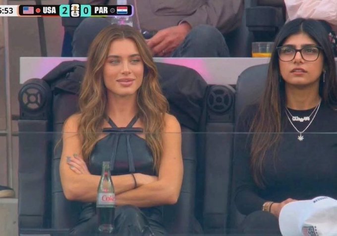 The image shows two women sitting next to each other, both wearing glasses. They are seated in a stadium or arena setting with a large screen behind them displaying the World Cup. One of the women has her arms crossed over her chest while the other woman is looking at something off-camera. There is also a bottle and a cup visible on the table between them. The post about RiverdotInc, which is related to the World Cup, suggests that they might be attending an event or watching a match together.