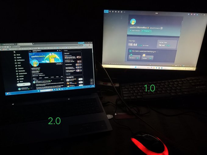 A dark workspace setup showing a laptop displaying X/Twitter labeled '2.0' and a monitor showing what appears to be a 'sashimi/MemeX' page labeled '1.0', with a red-glowing mouse in the foreground.