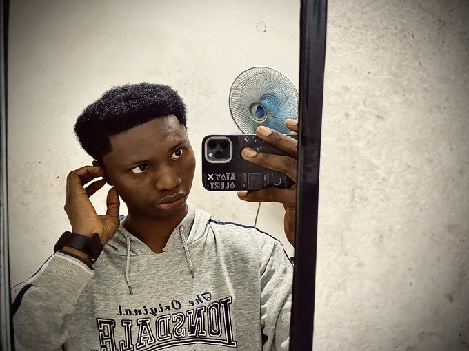 A mirror selfie of a young man wearing a gray 'Long Beach Original' hoodie, holding up a black phone case with 'STAY' and 'ALOST' text while touching his ear.
