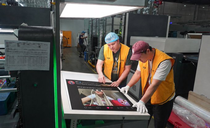Two workers in yellow VIP vests and hairnets inspect a printed Razer advertisement featuring 'Digital World Headset' text and a '1.7K' view count on a large flatbed printer in a workshop.