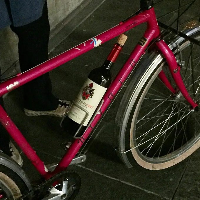 A pink bicycle carries a wine bottle labeled 'CAPOFOSCO' in its frame rack at night.