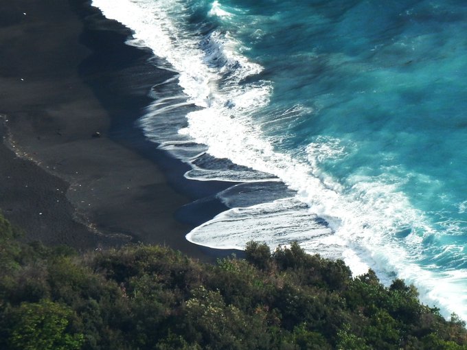 An aerial view shows turquoise ocean waves crashing onto a dark black sand beach, with lush green vegetation visible along the shoreline.